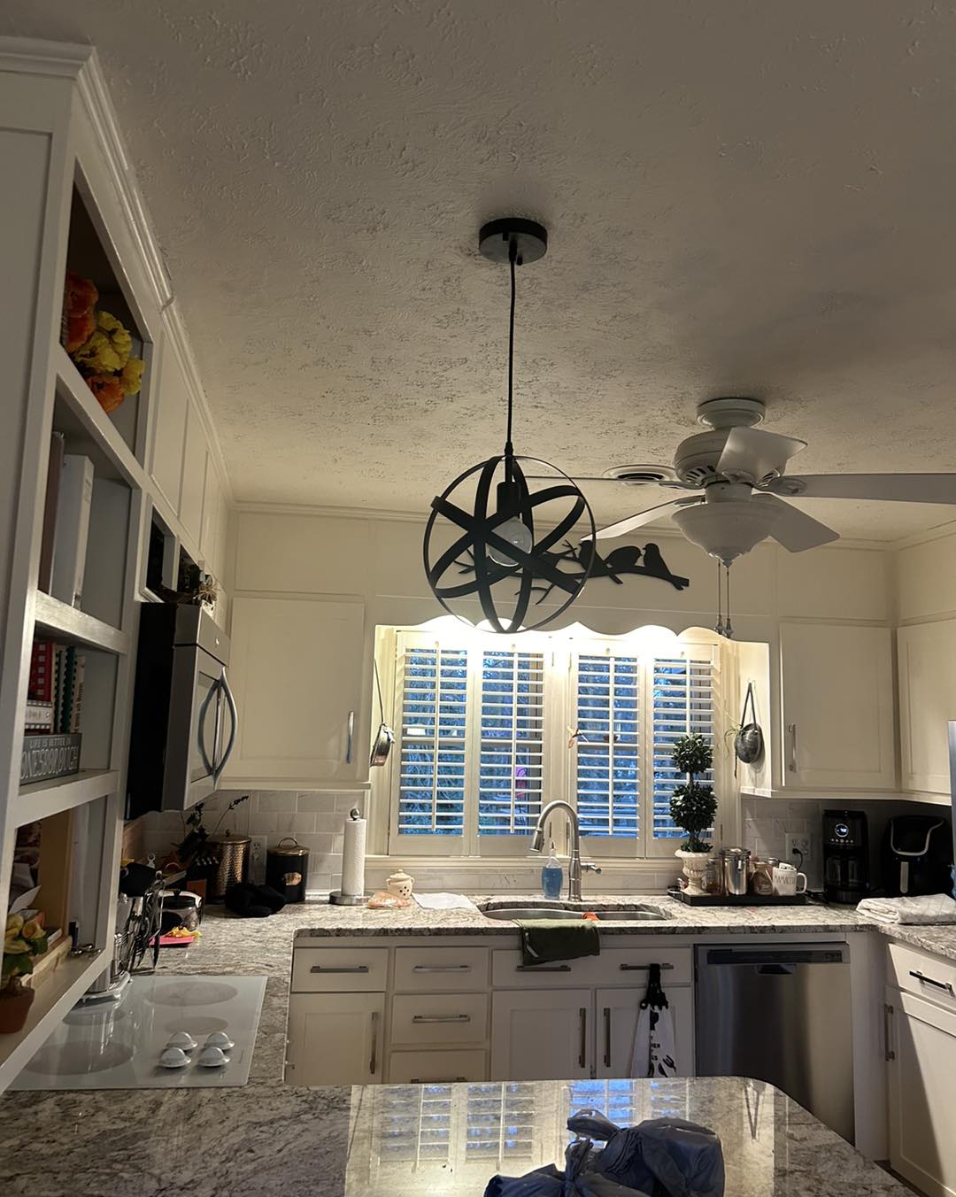 A white kitchen with a globe-style pendant light, granite countertops, a sink below a window with shutters, and various kitchen appliances and decor.