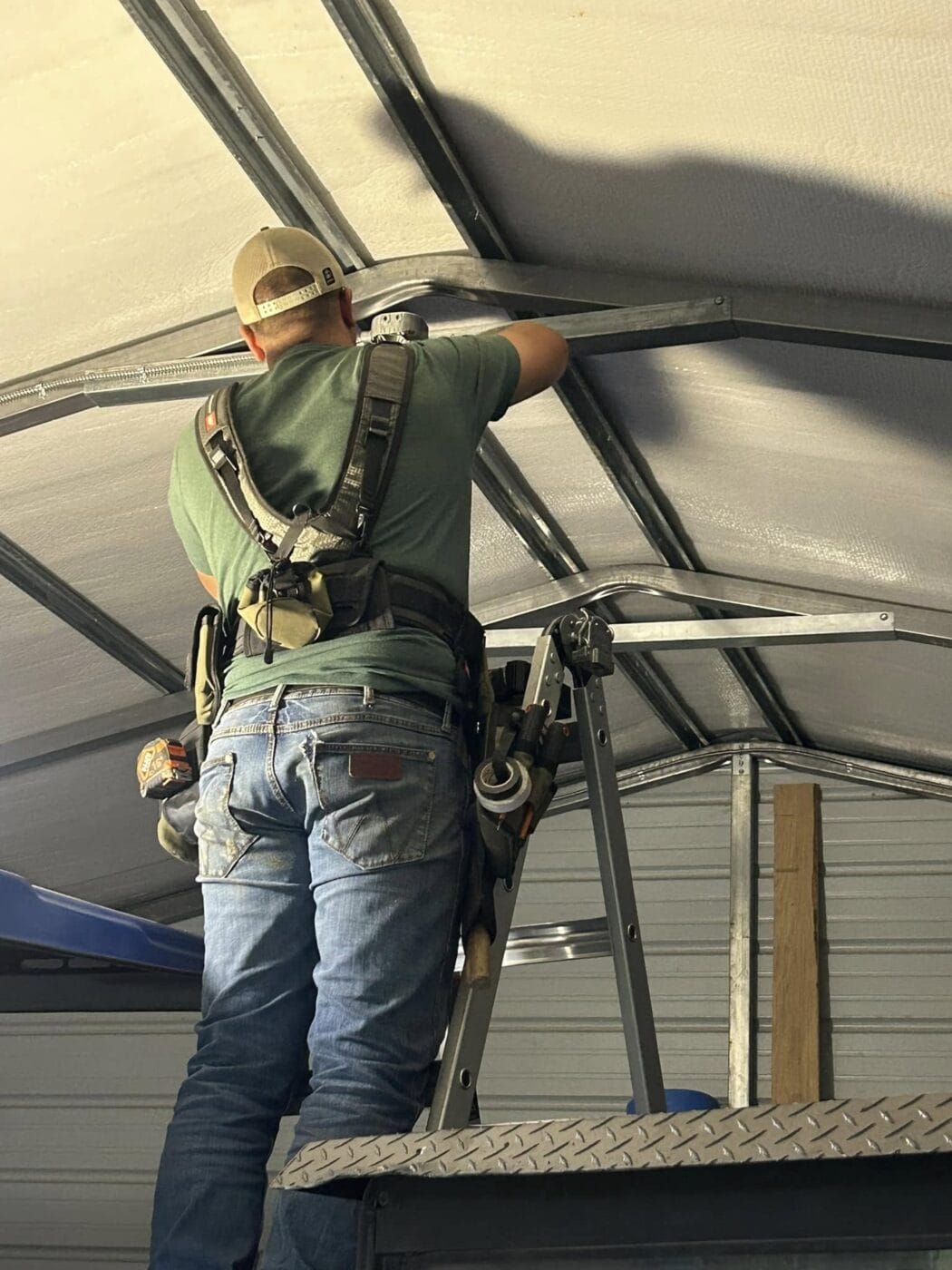 Person in a green shirt and jeans standing on a ladder, using a tool to install or repair a metal structure inside a building.
