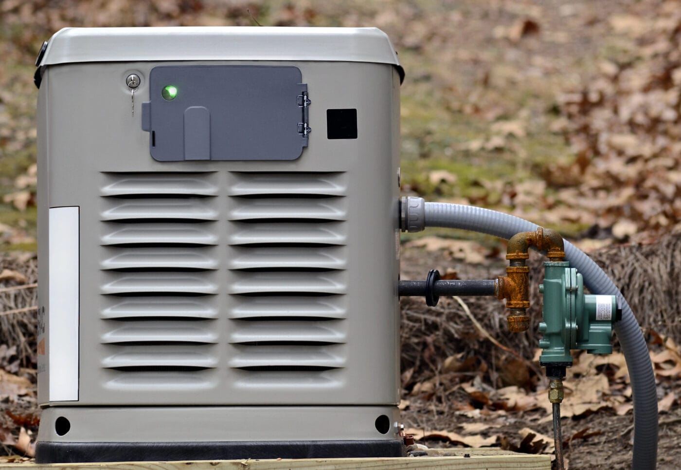 A gray metal generator with a vented front, attached to a green regulator and pipes, positioned outdoors on a wooden platform, with dry leaves scattered around.