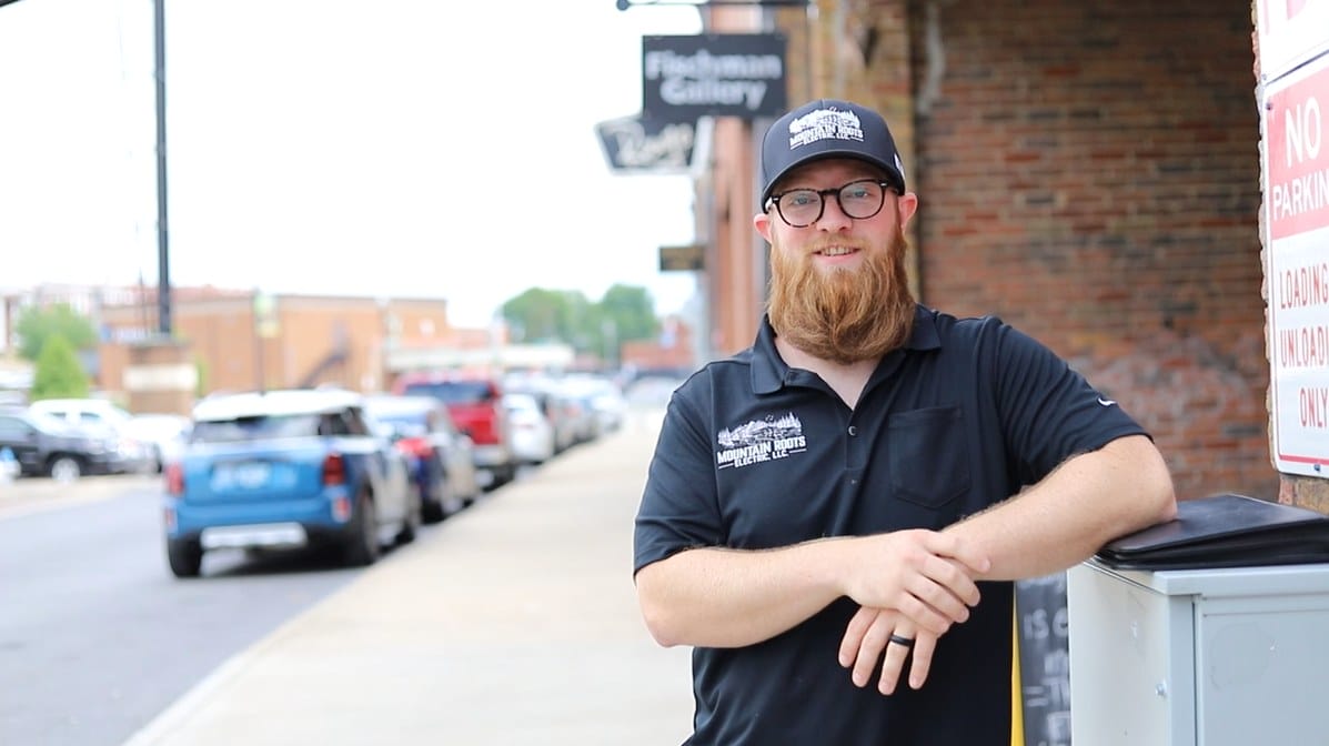 A bearded man in a black shirt and cap leans against a post on a city sidewalk. Cars are parked along the street.