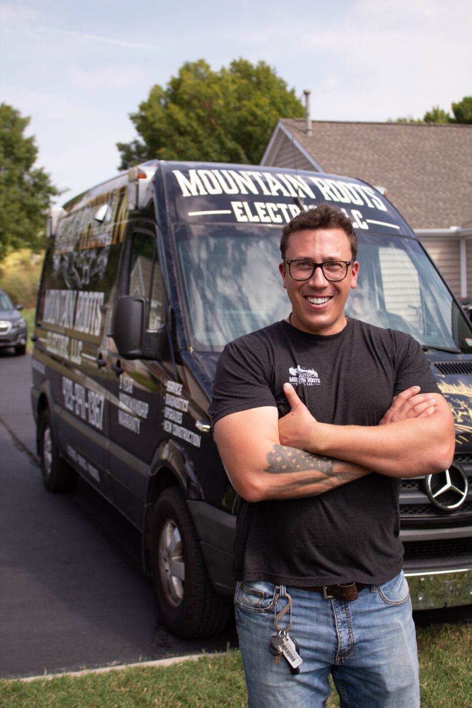 Man in glasses and black t-shirt smiling with arms crossed, standing in front of a black van labeled Mountain Roots Electric.