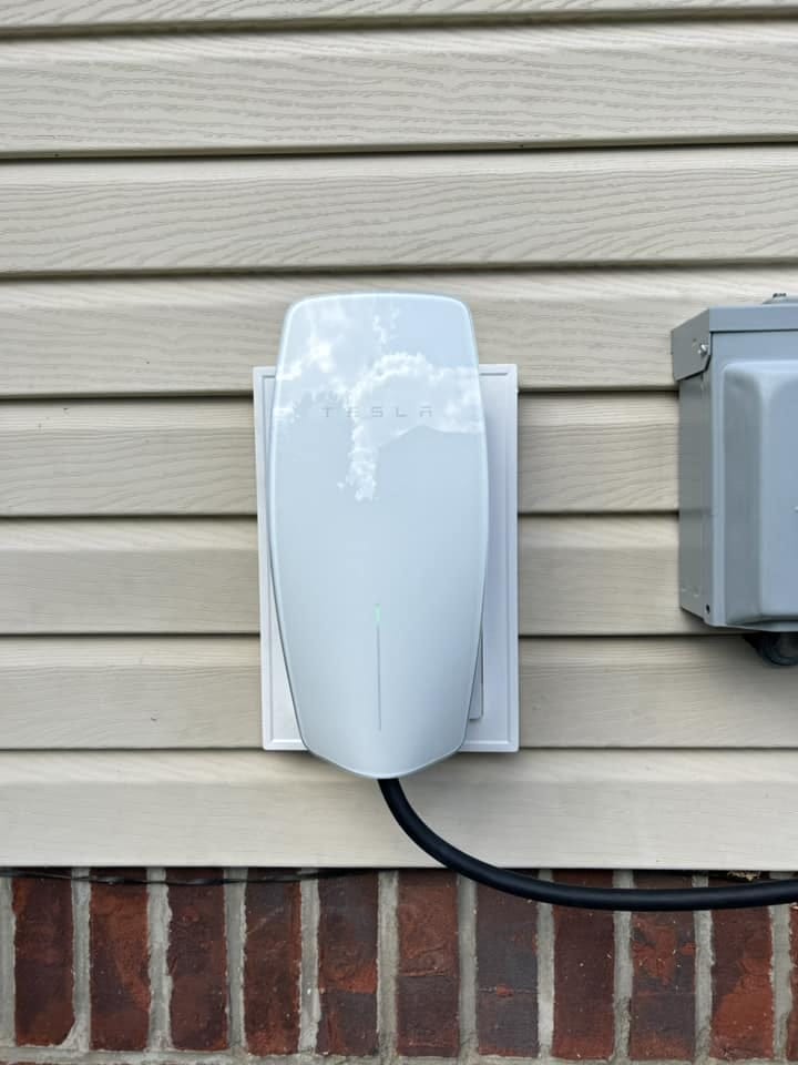 A white Tesla wall charger is mounted on a beige siding wall with visible power lines connected.